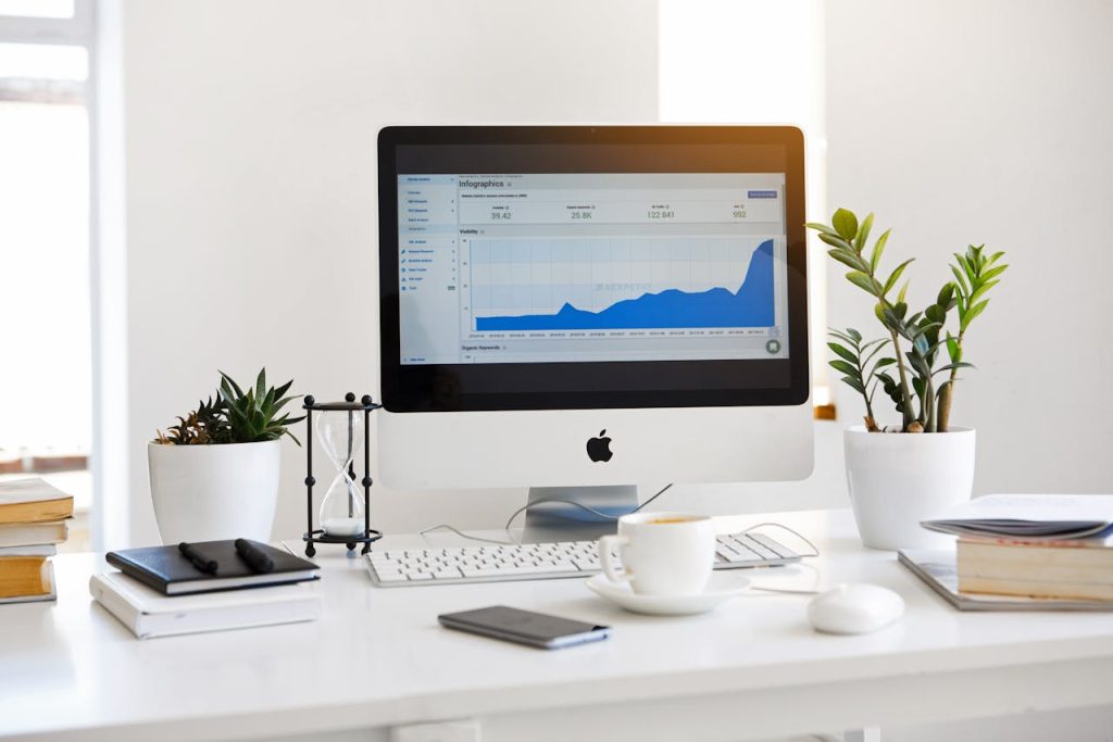 silver-imac-displaying-line-graph-placed-on-desk-572056 A clean, contemporary workspace featuring a desktop with analytics on the screen and plants for a fresh look.