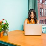 Home Confident woman in a bright office working on a laptop, surrounded by urban scenery and greenery.